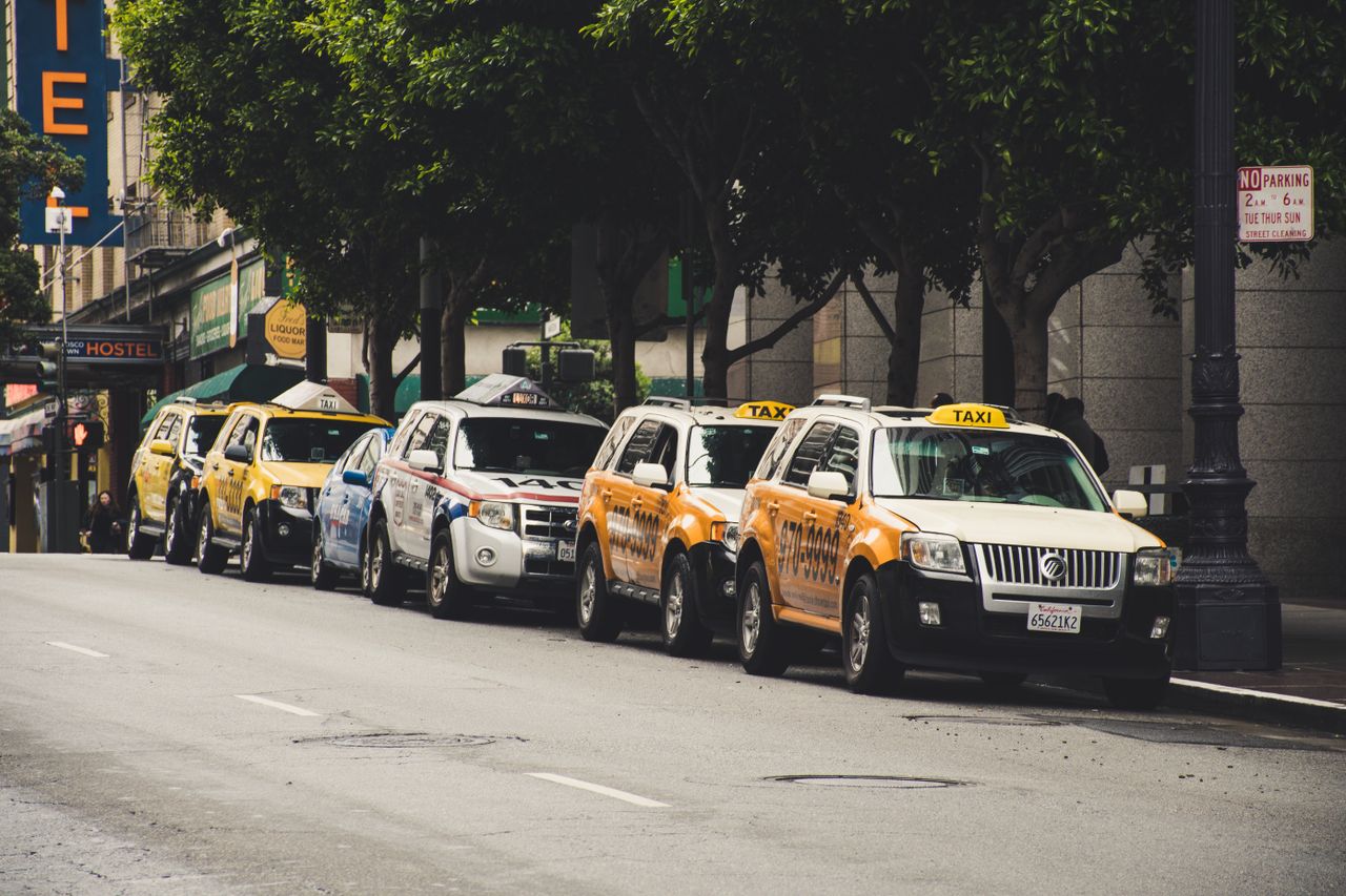 about-img A row of taxis parked on a city street with trees and urban background.
