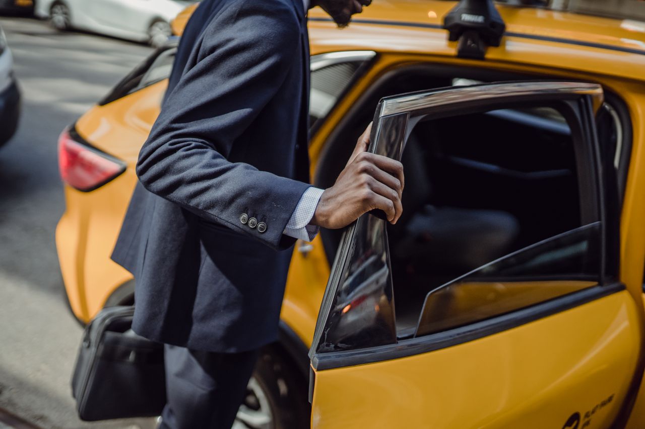 ours-journey A businessman in a suit opens the door to a yellow taxi on a city street during the day.